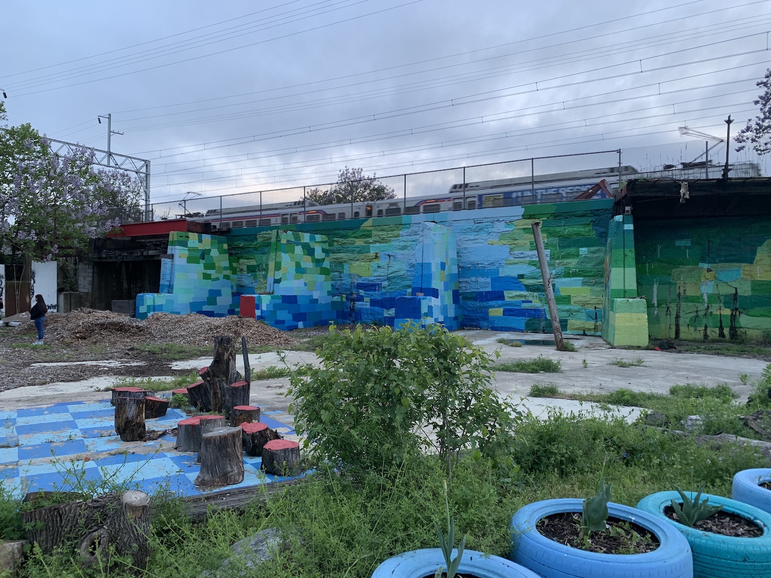 Image description: urban farm with painted ball courts & train in the background. in the foreground are tires painted blue with plants inside them. There is an oversized checkerboard in the center with blue squares and tree stumps as pieces. In the back to the left is a person standing by some mulch. There is a blue & green patterned mural on the wall in the back with an idled train atop the wall.